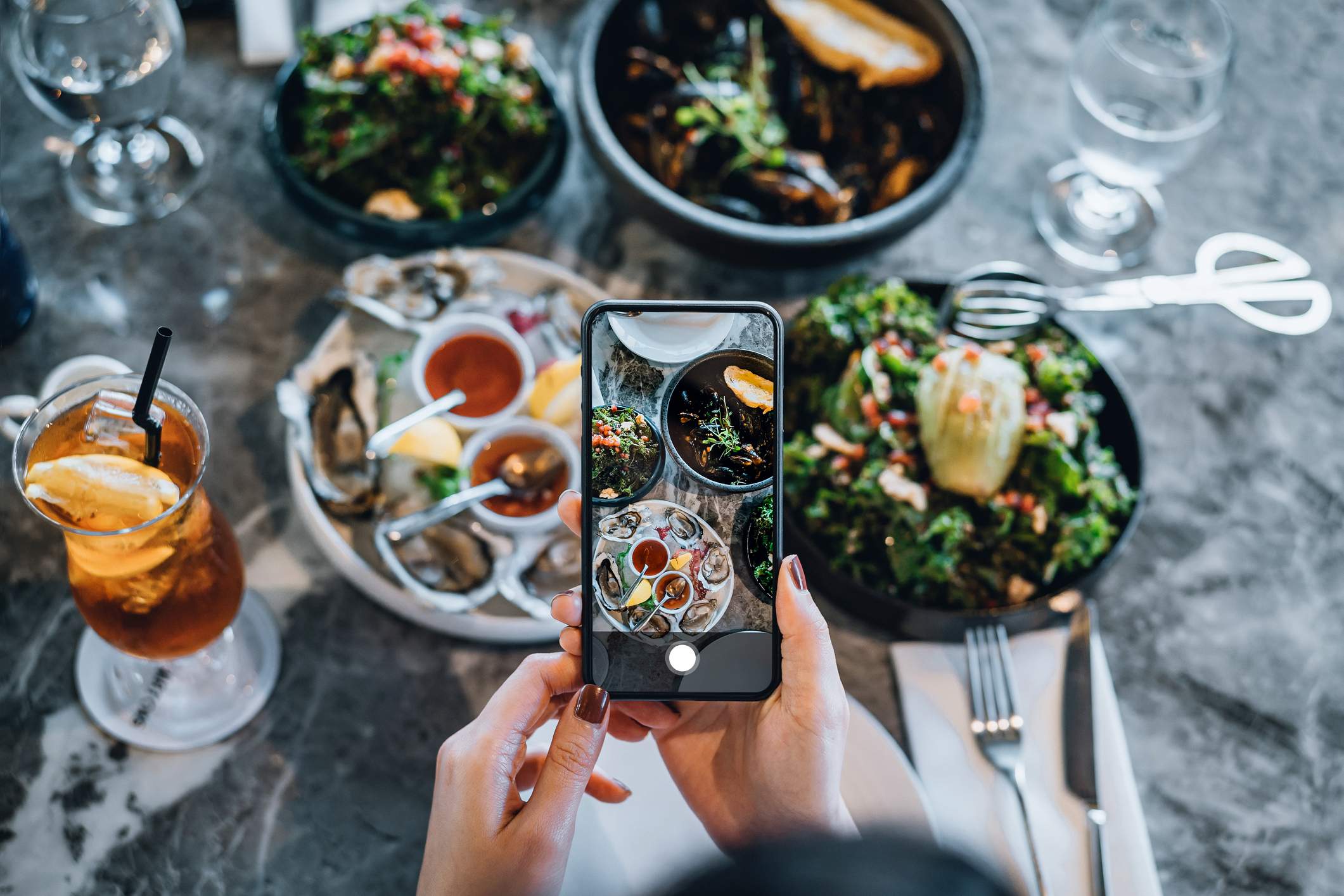 Image depicts a person out of frame holding a phone to take a photo of food on a table. 