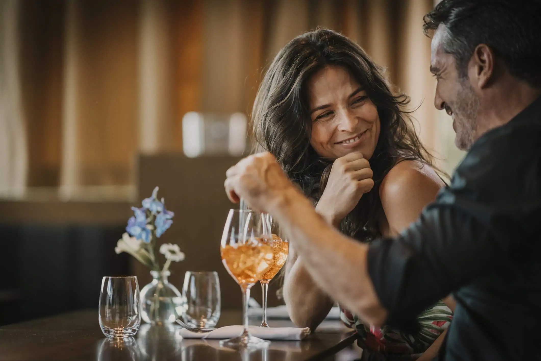 Image depicts two people sitting at a table at a restaurant. They are drinking orange drinks in wine glasses. One of the people has turned back to smile at the person to their left. 