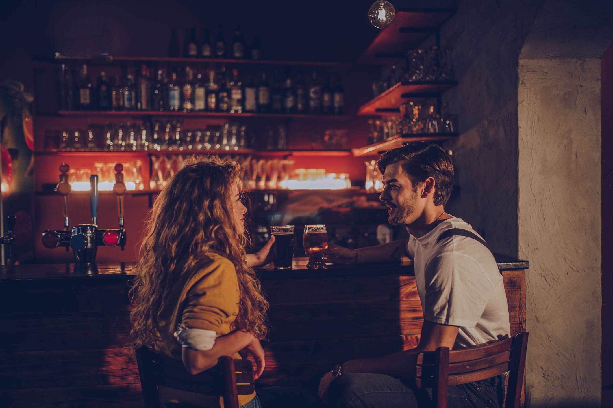 Image depicts two people sitting at a bar. They are each having a beer. One is wearing a white short sleeve shirt and the other is wearing a yellow button up. The restaurant is dimly lit. 