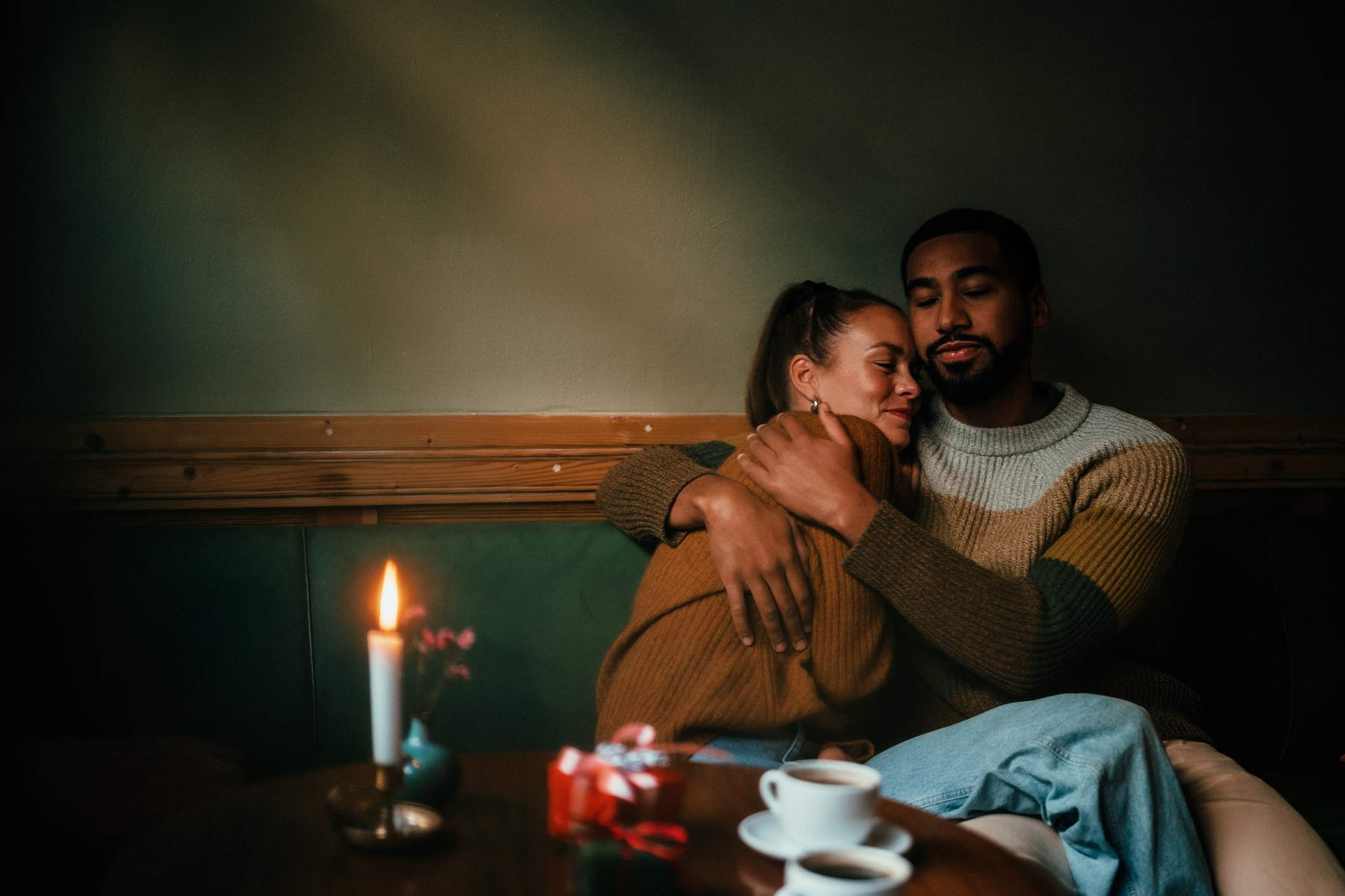 Image depicts a couple snuggling in a restaurant booth. There are two small gifts and coffee on the table in front of them. 