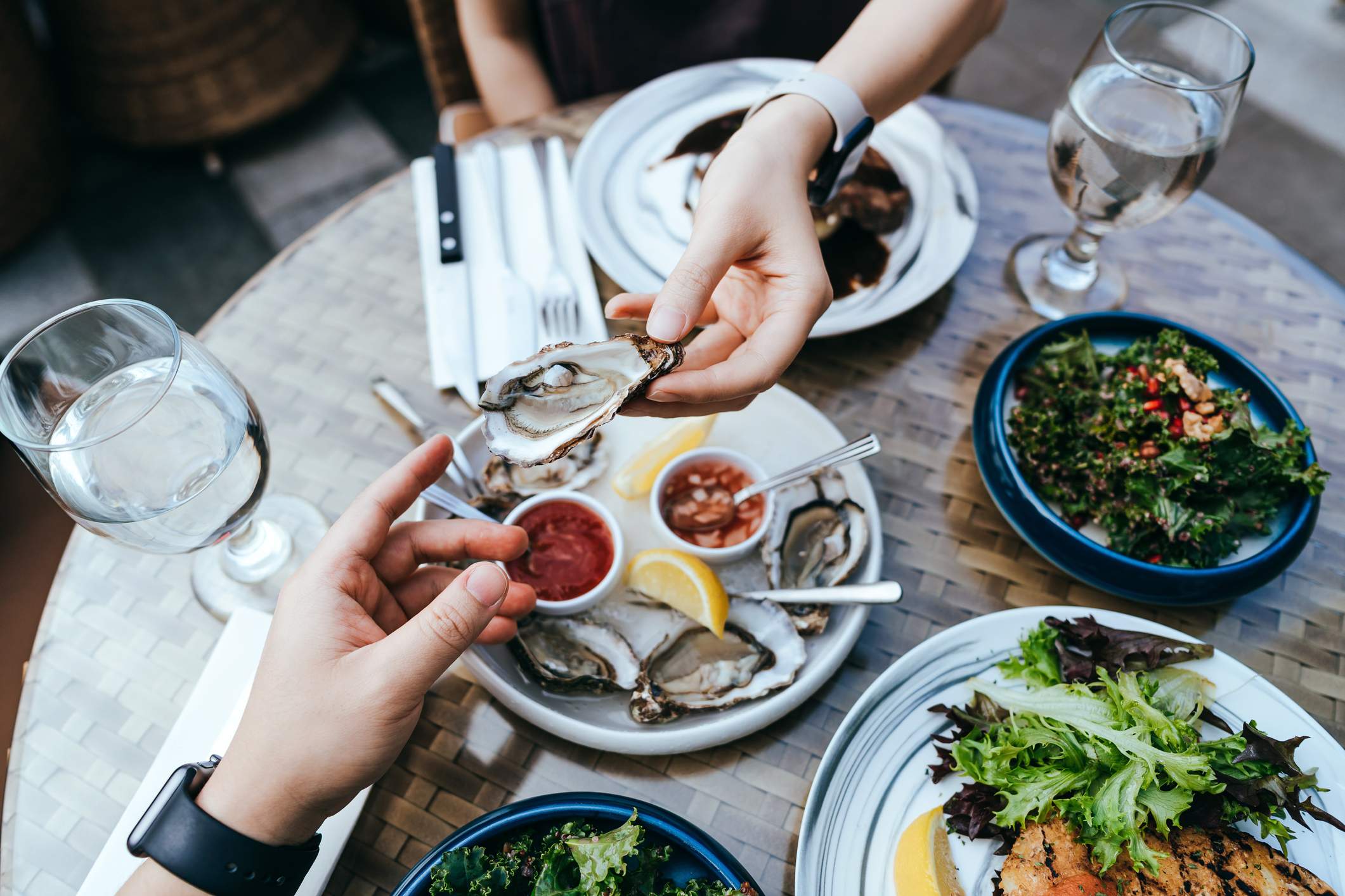 Image depicts two people sitting at a restaurant sharing oysters. There are other plates of food on the table. 