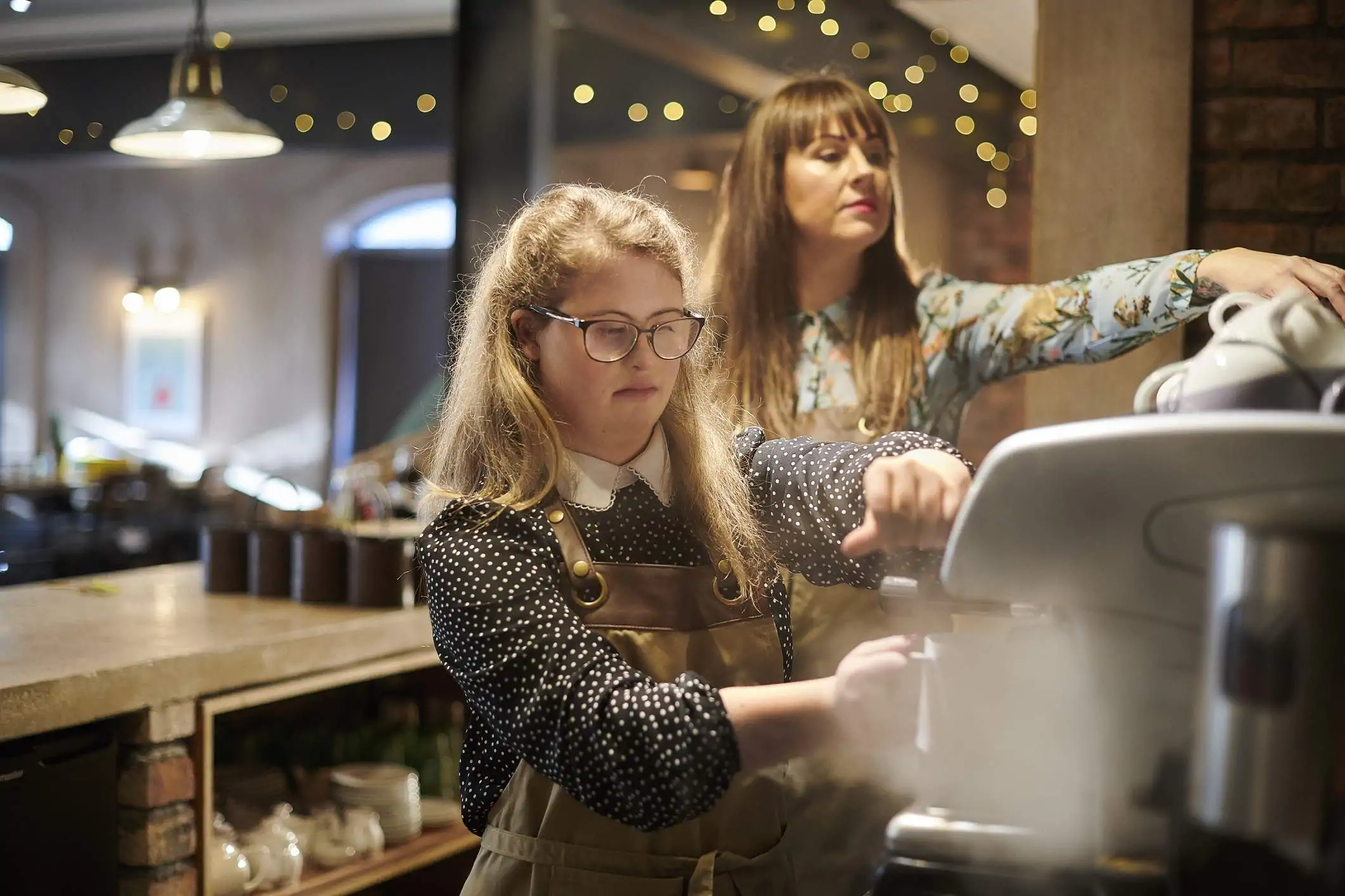 Image depicts a young worker pouring a cup of coffee while a supervisor is working in the back.