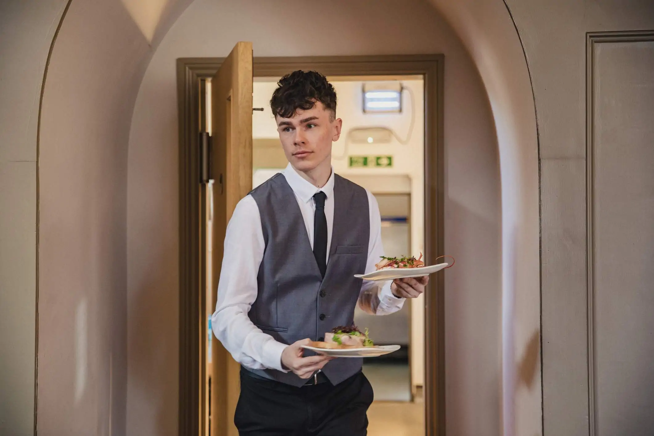 Image depicts a young restaurant worker bringing plates of food out to guests.