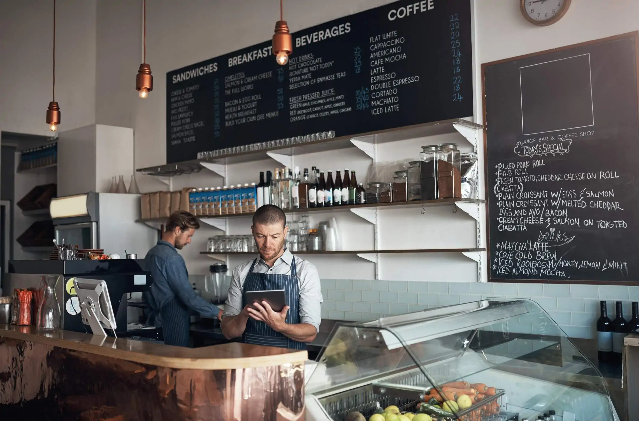 Image depicts two restaurant workers behind a counter. One is working on a tablet.