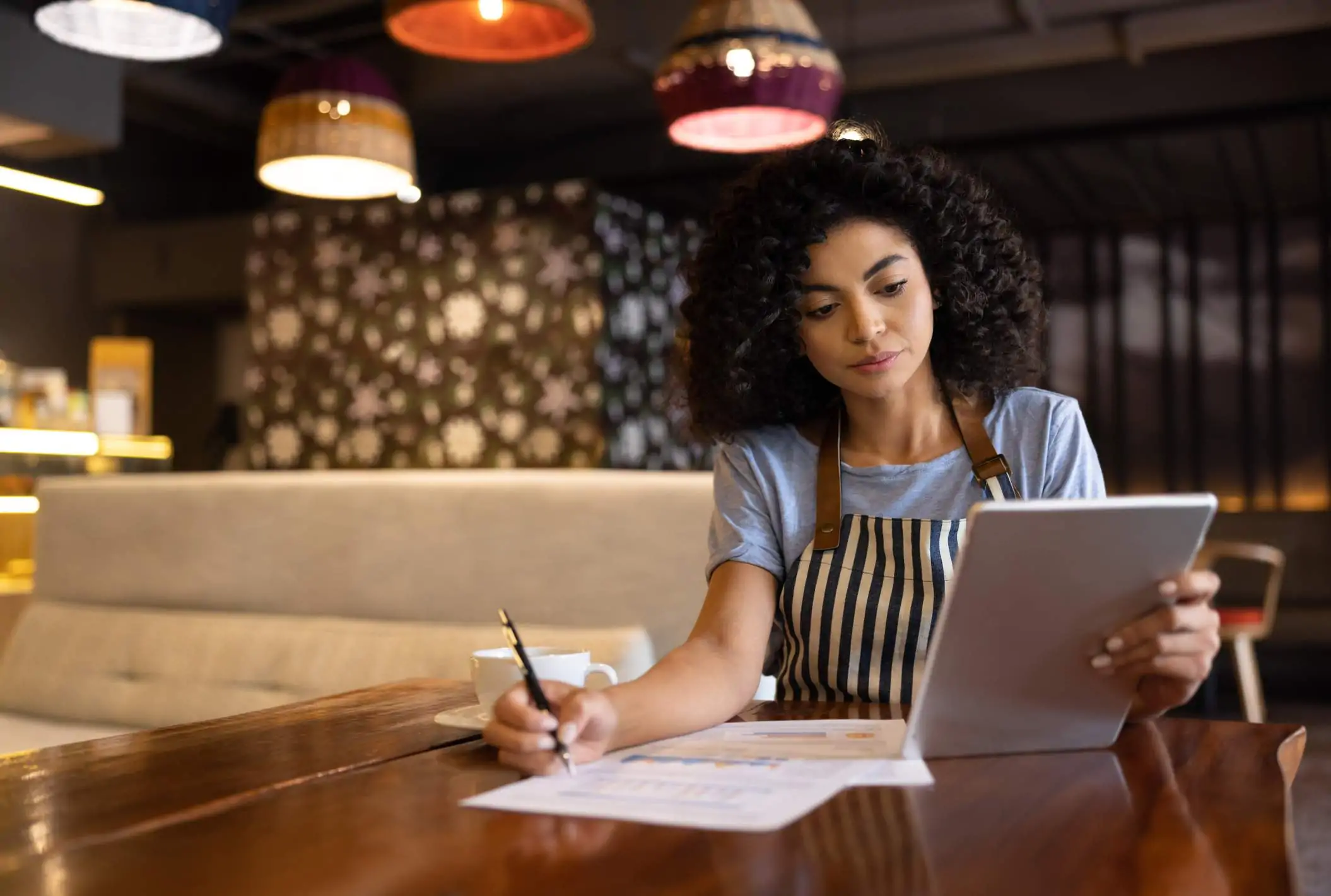 Image depicts a person in a striped apron looking at a tablet while writing on a piece of paper.