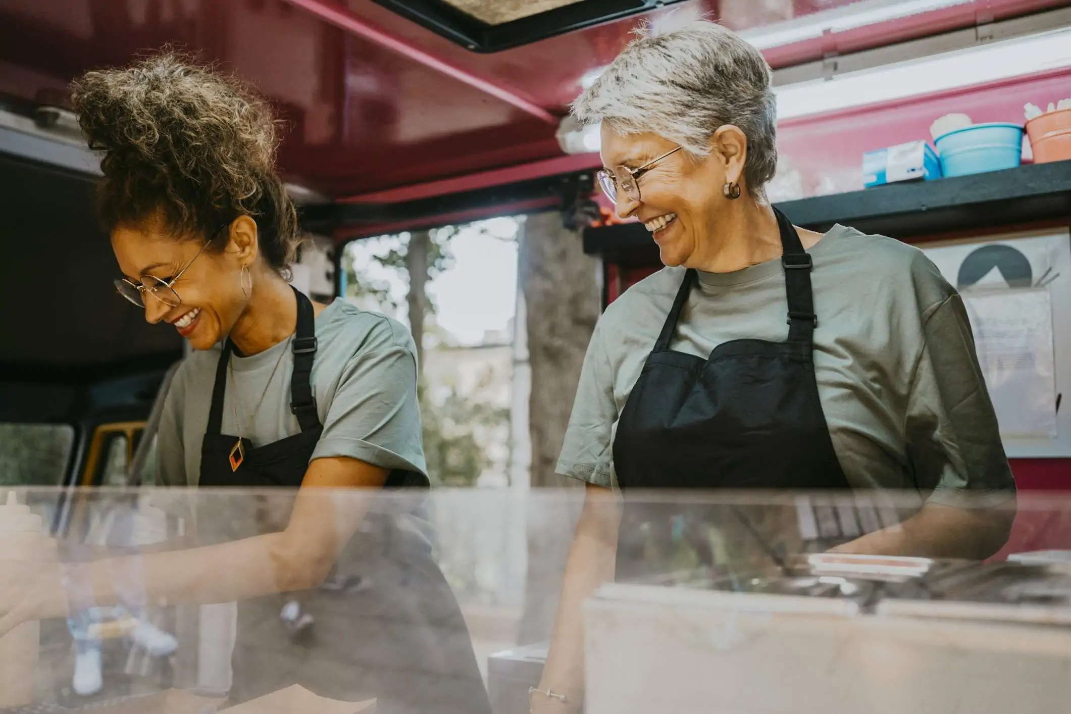 Image depicts two people wearing teal shirts and black aprons working inside of a food truck.