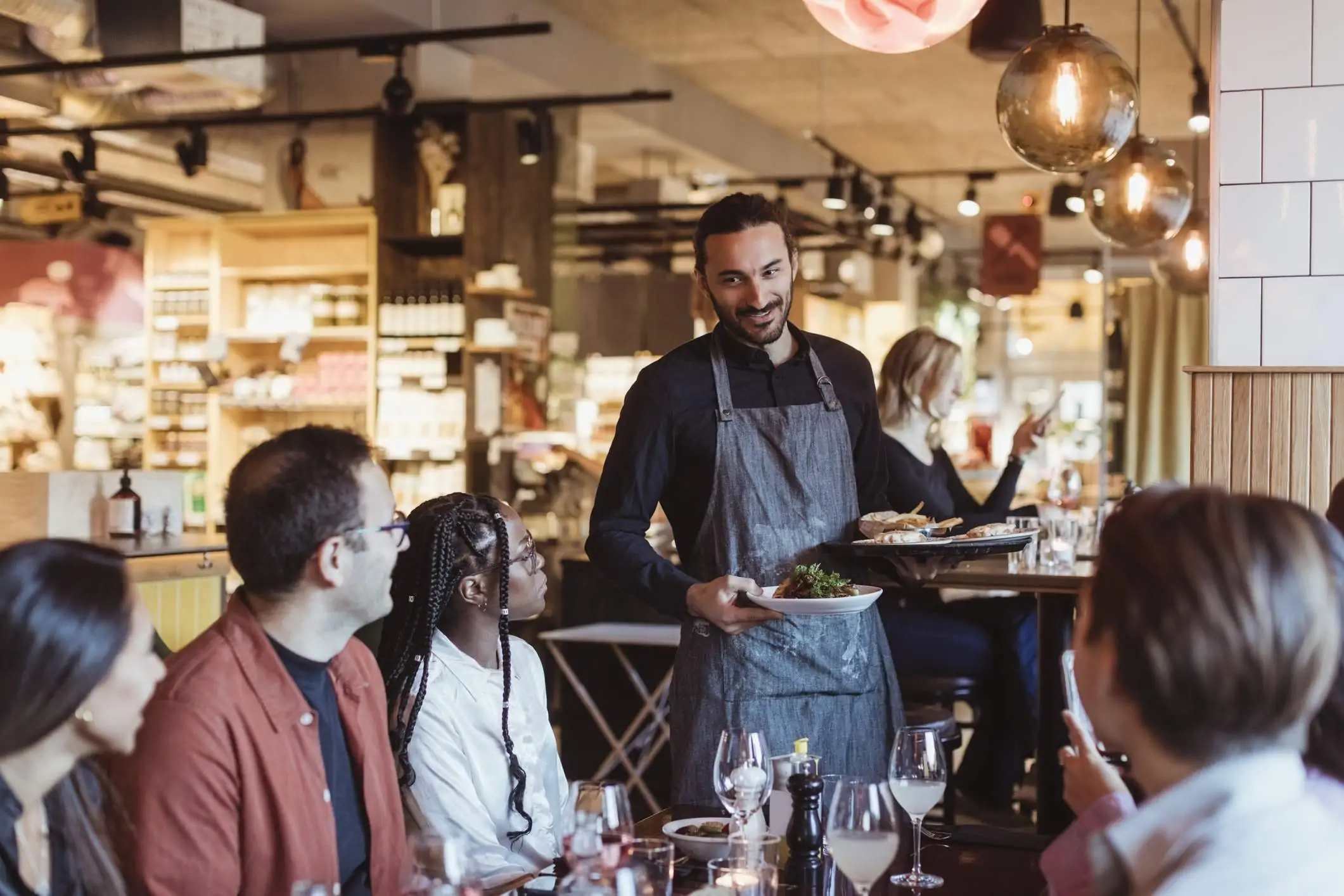 Image depicts a server in a gray apron delivering two plates of food to a group seated at a large table.