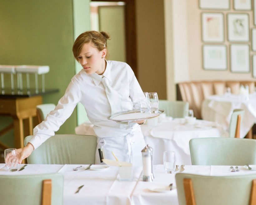 Image depicts a server setting a table. They are dressed in all white and the chairs in the restaurant are a mint green hue.