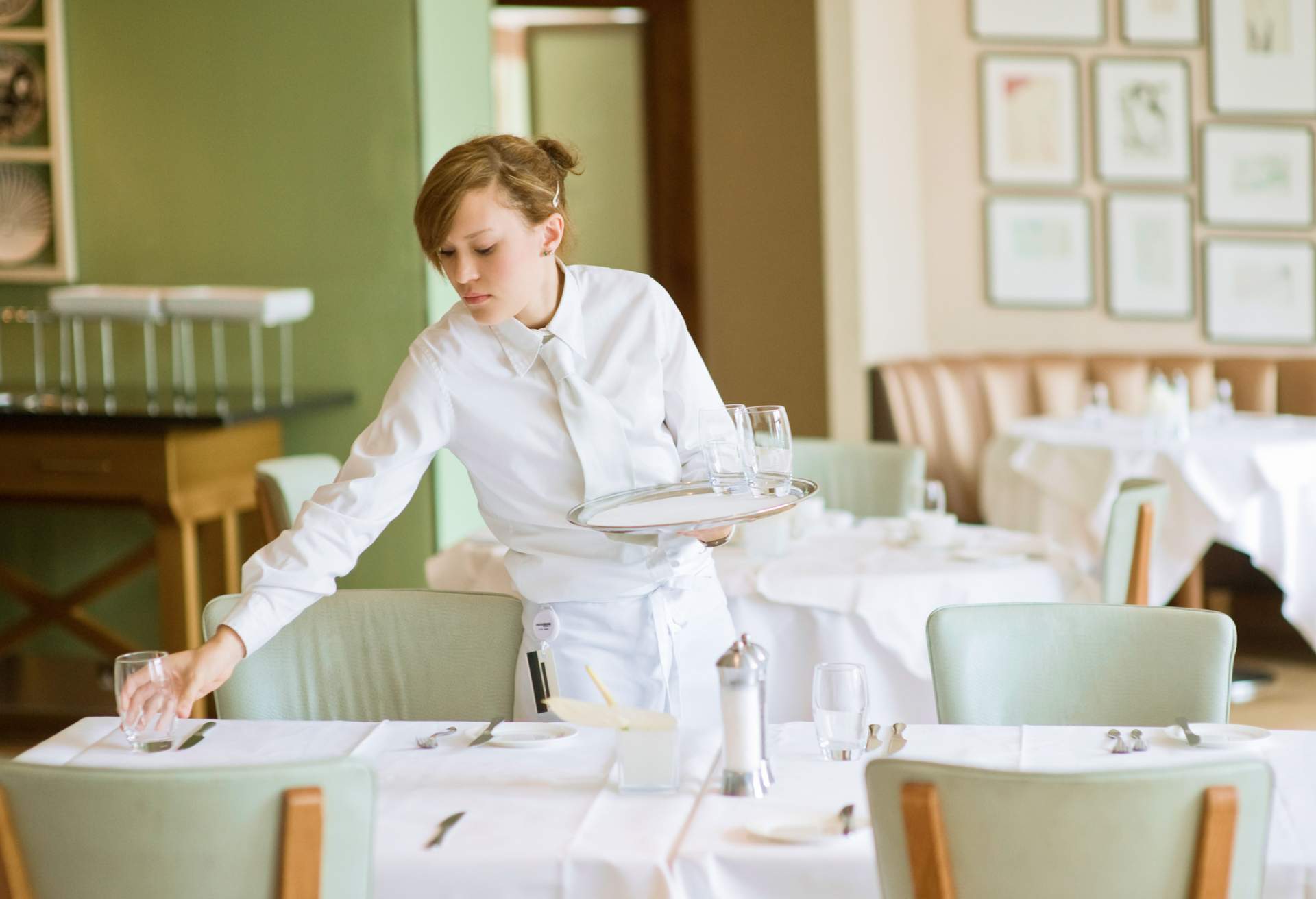 Image depicts a server setting a table. They are dressed in all white and the chairs in the restaurant are a mint green hue.