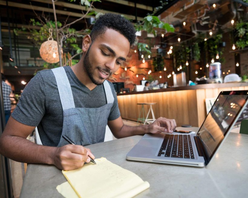 Image depicts a restaurant worker writing on a pad of paper while working on a computer. They are seated at a table in a restaurant.