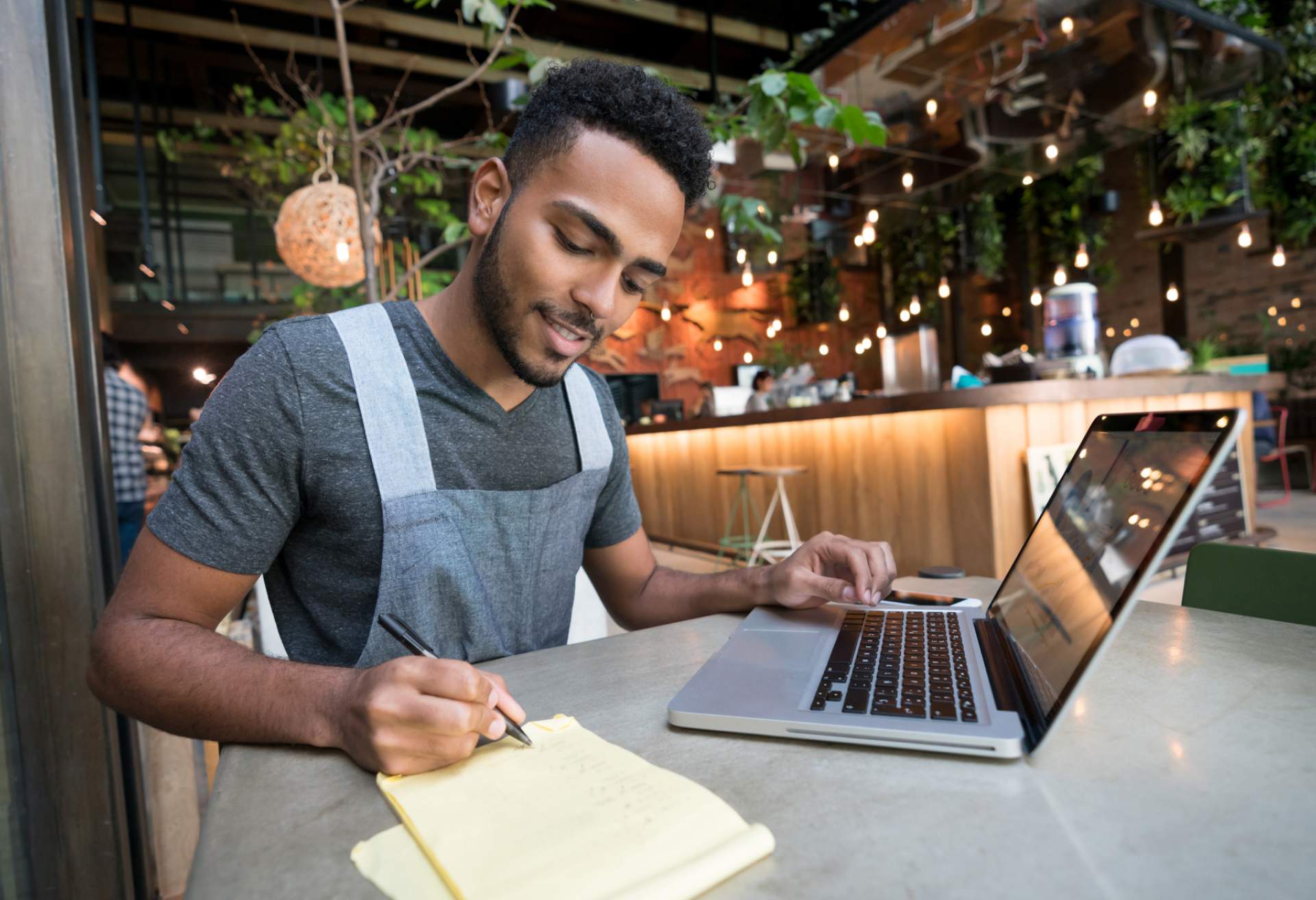 Image depicts a restaurant worker writing on a pad of paper while working on a computer. They are seated at a table in a restaurant.