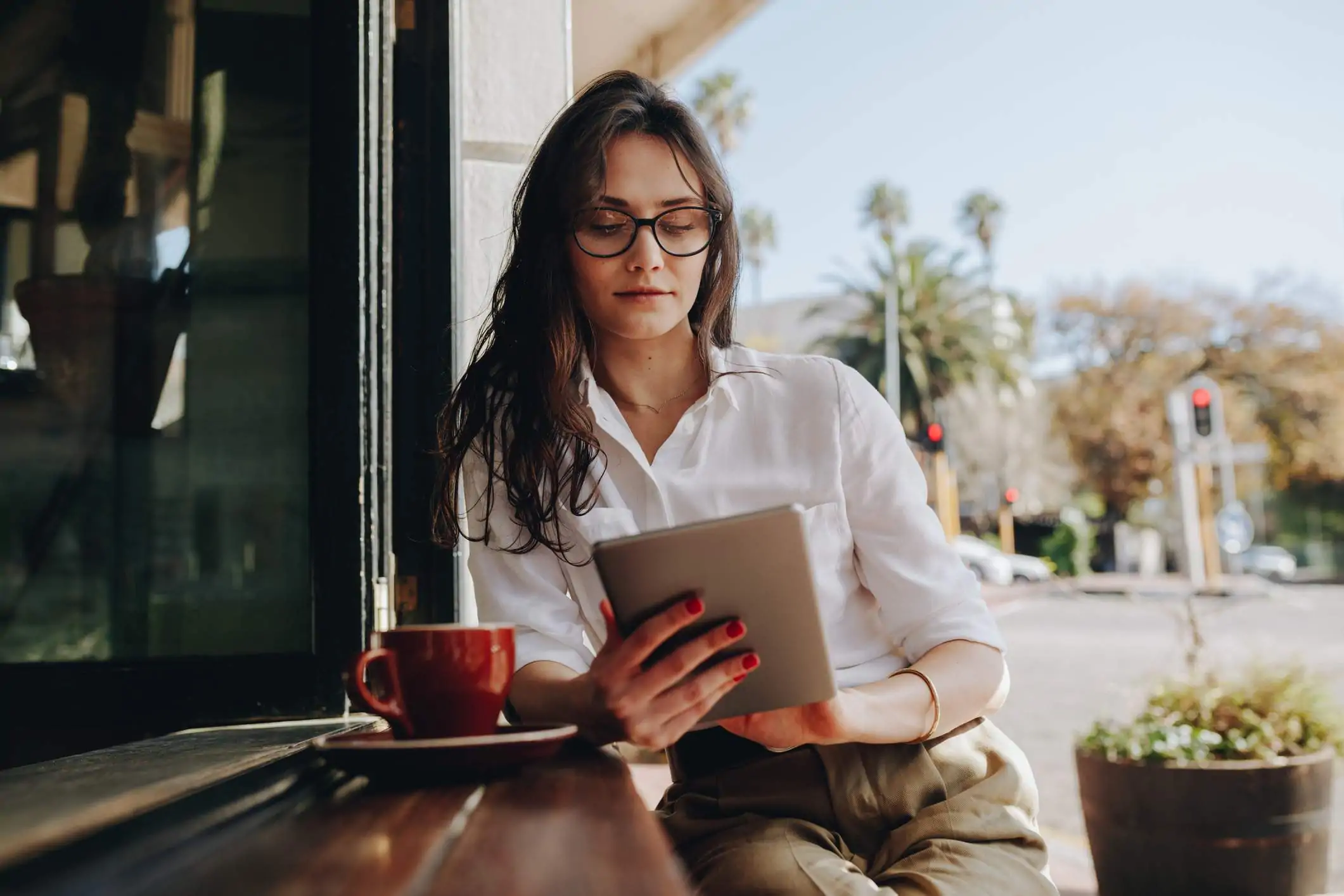 A restaurant worker wearing glasses is using a tablet while sitting at an outside counter of the restaurant.