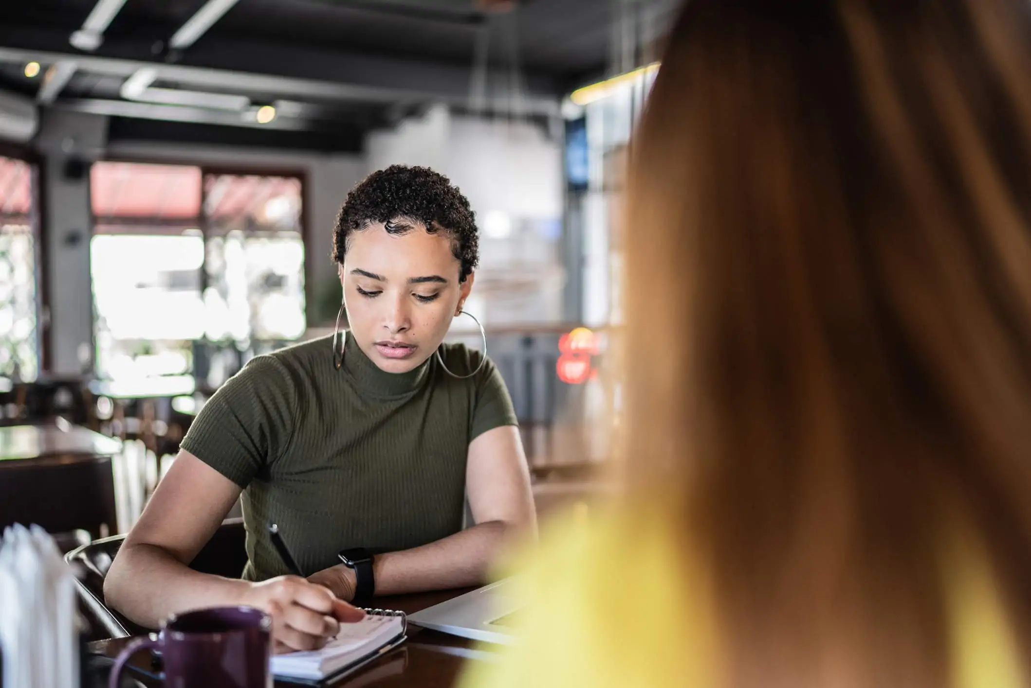 Image depicts a person interviewing another person at a restaurant. The interviewee is taking notes.