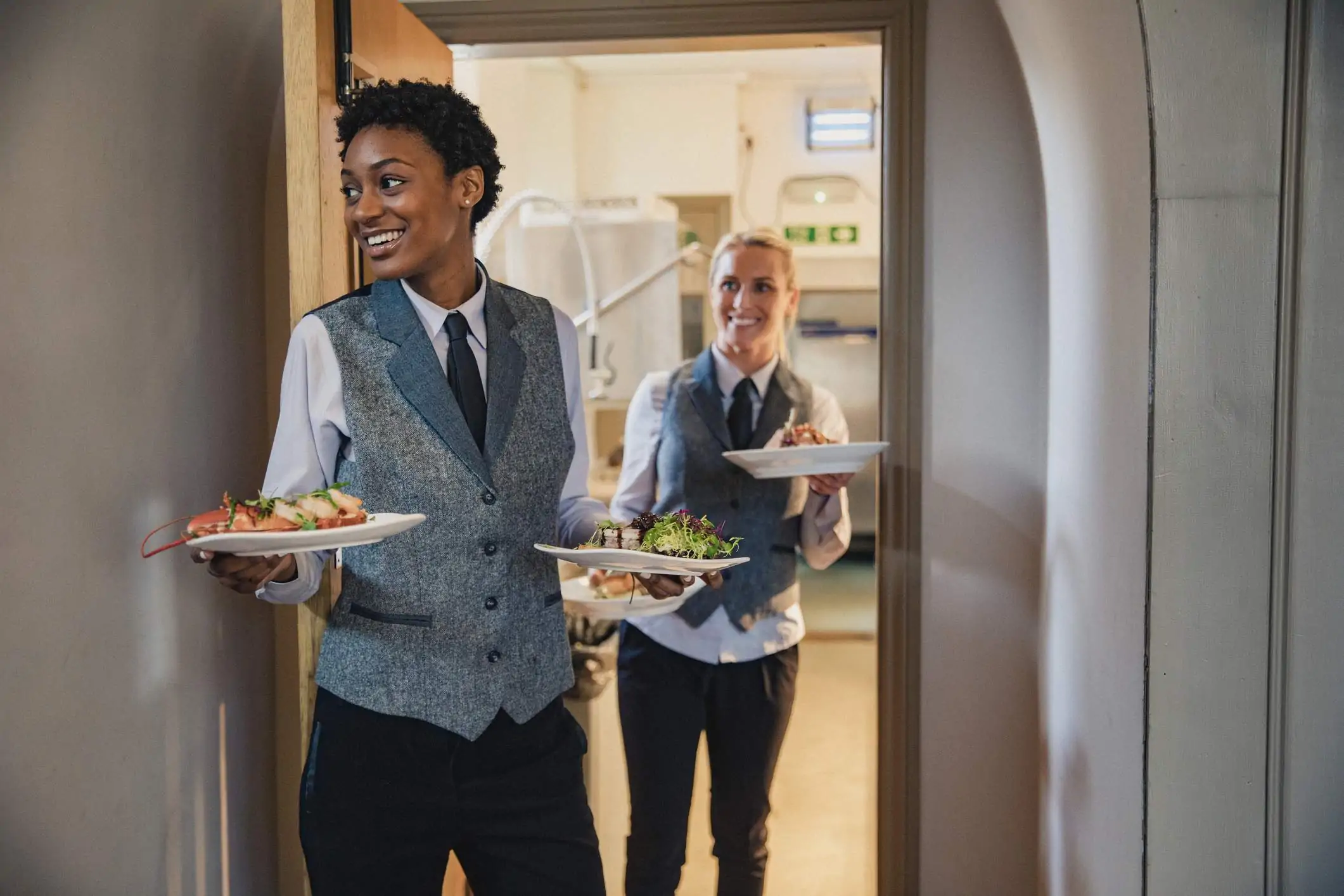 Image depicts two servers walking through a doorway as they carry plates of food.