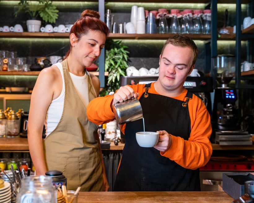 Image depicts a young barista pouring a coffee as a supervisor watches.
