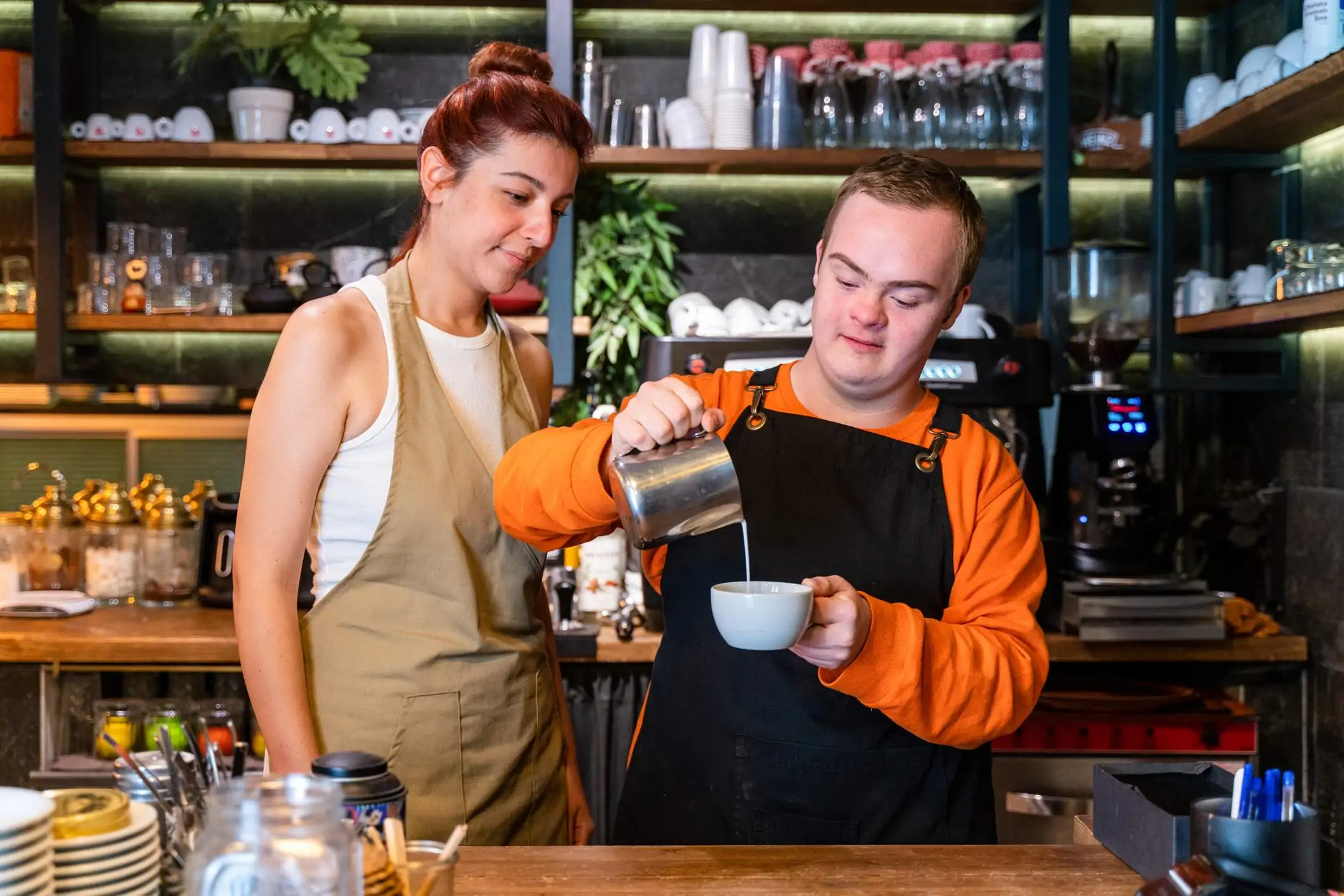 Image depicts a young barista pouring a coffee as a supervisor watches.