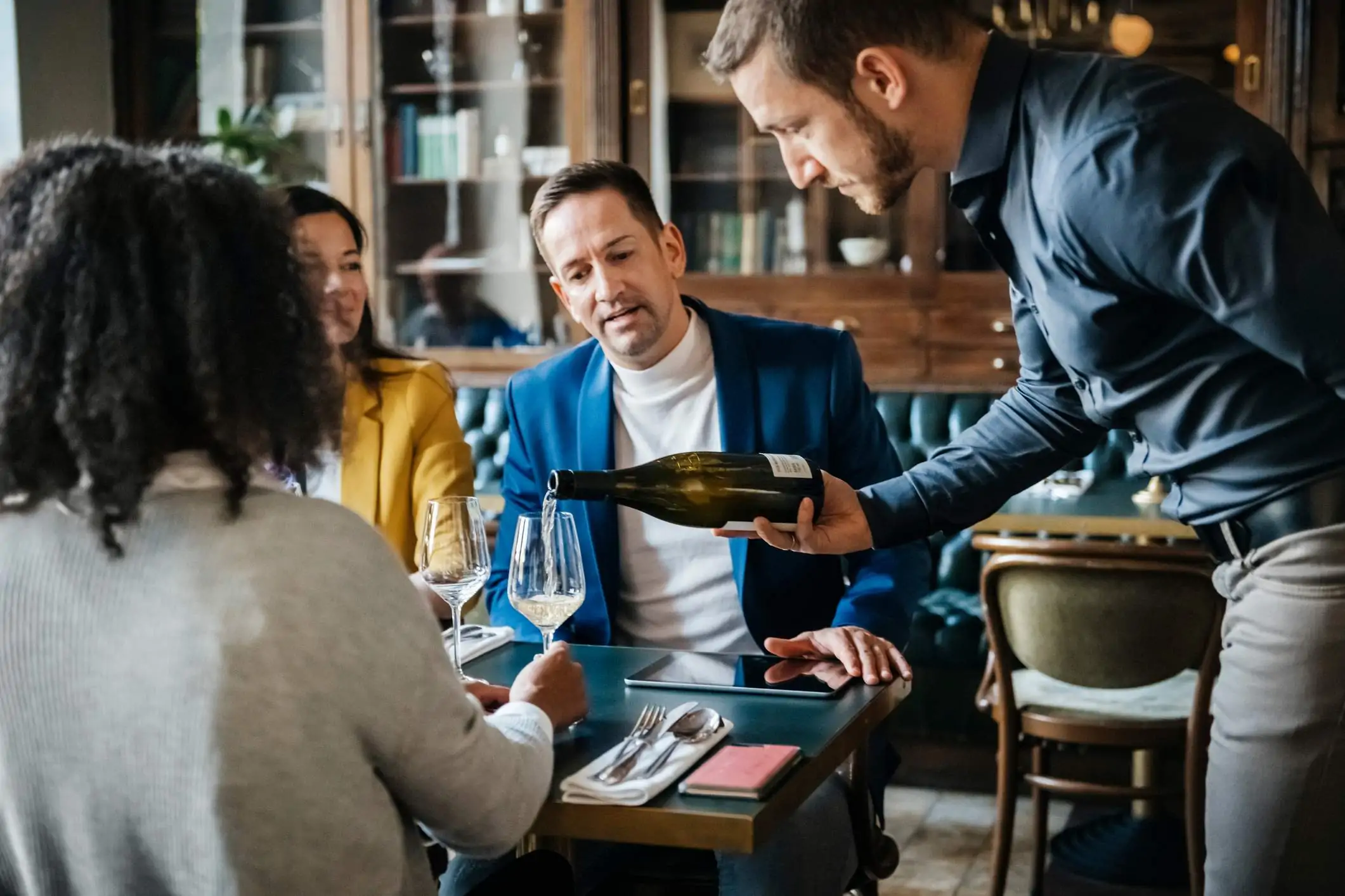 Image depicts a group of people dining at a restaurant and a server pouring some wine.